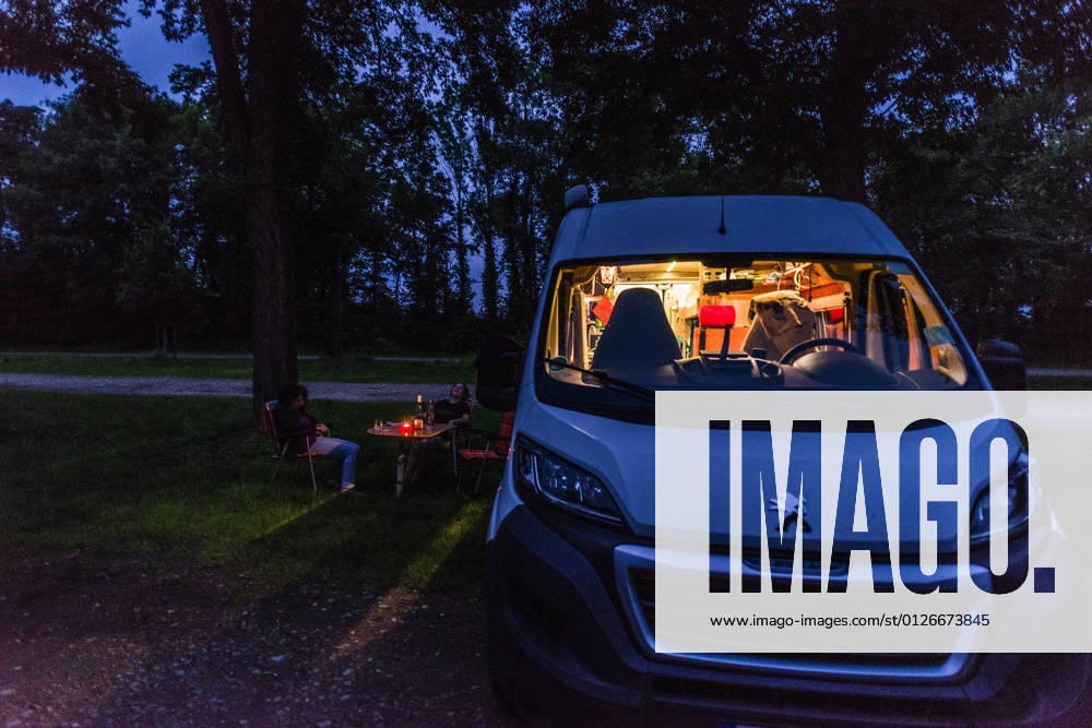 Camper sitzen zur blauen Stunde an einem lauen Sommerabend vor ihrem Camper auf Campingstühlen. Baden Württemberg, Deutschland. Mit einem Campervan unterwegs
