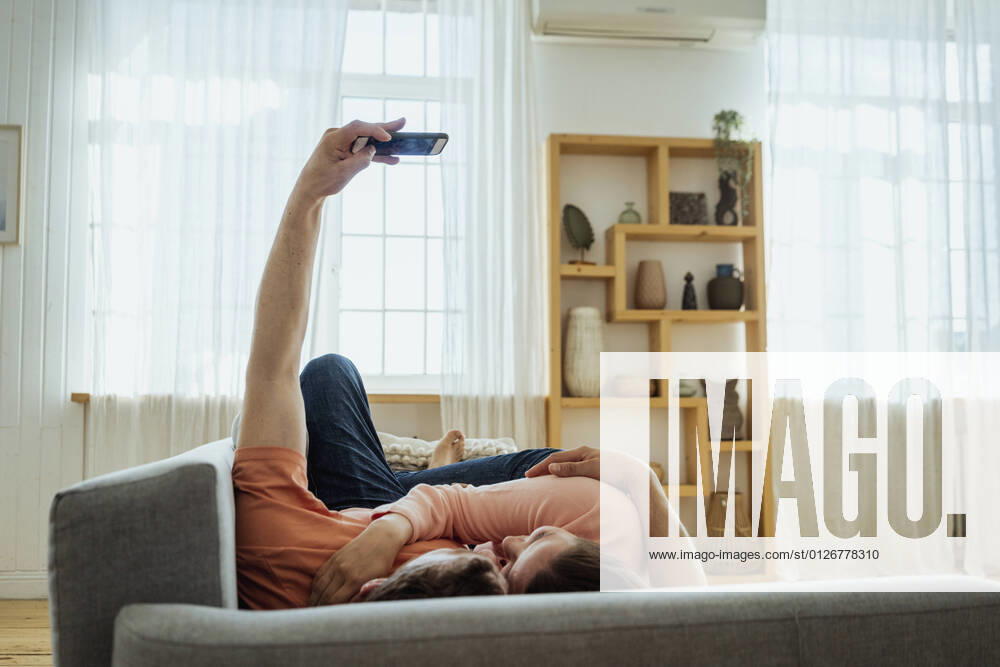 IMAGO Creative: Jahreszeit Winter, Couching Man embracing woman while taking selfie through mobile phone at home model released Symbolfoto property released VPIF04363