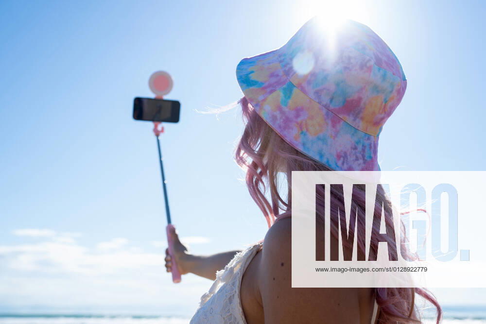 Unrecognizable female with pink hair and in summer outfit taking self shot on smartphone on selfie stick while standing on sunny day on seashore OscarDeDios_SummertimeLucia_11.jpg