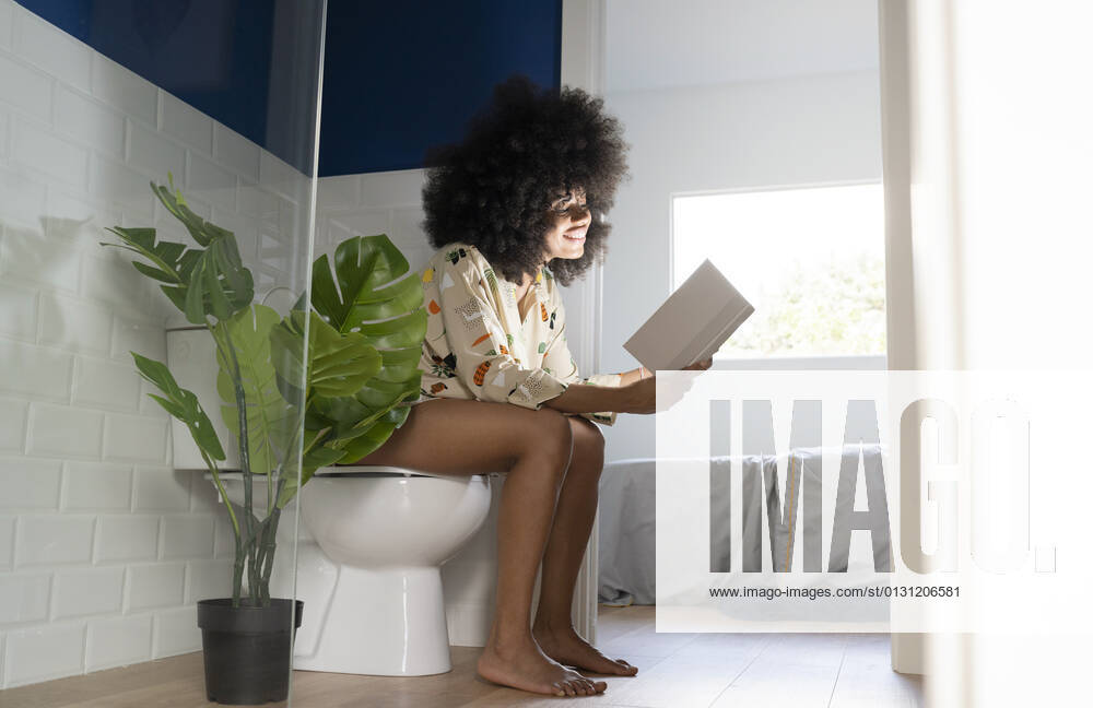 Afro woman reading book near potted plant while sitting on toilet seat at home model released Symbolfoto property released JCCMF03266