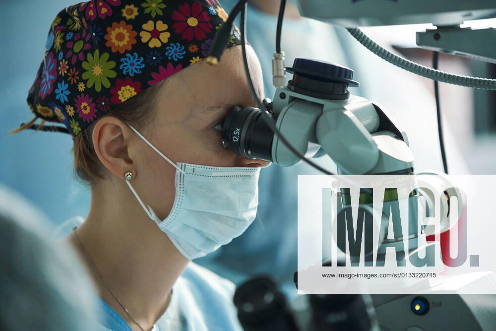 Adult female doctor in sterile mask and ornamental medical cap looking through surgical microscope against crop coworker in hospital, Model released SergeyMironov_Surgery_49.jpg