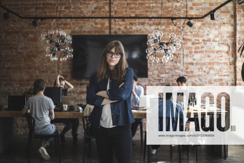 Female entrepreneur standing with arms crossed while colleagues working in creative office Sweden, Stockholm, model released, property released
