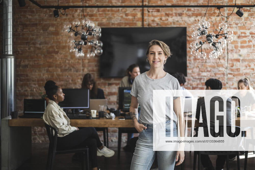 Female entrepreneur standing with arms crossed while colleagues working in creative office Sweden, Stockholm, model released, property released