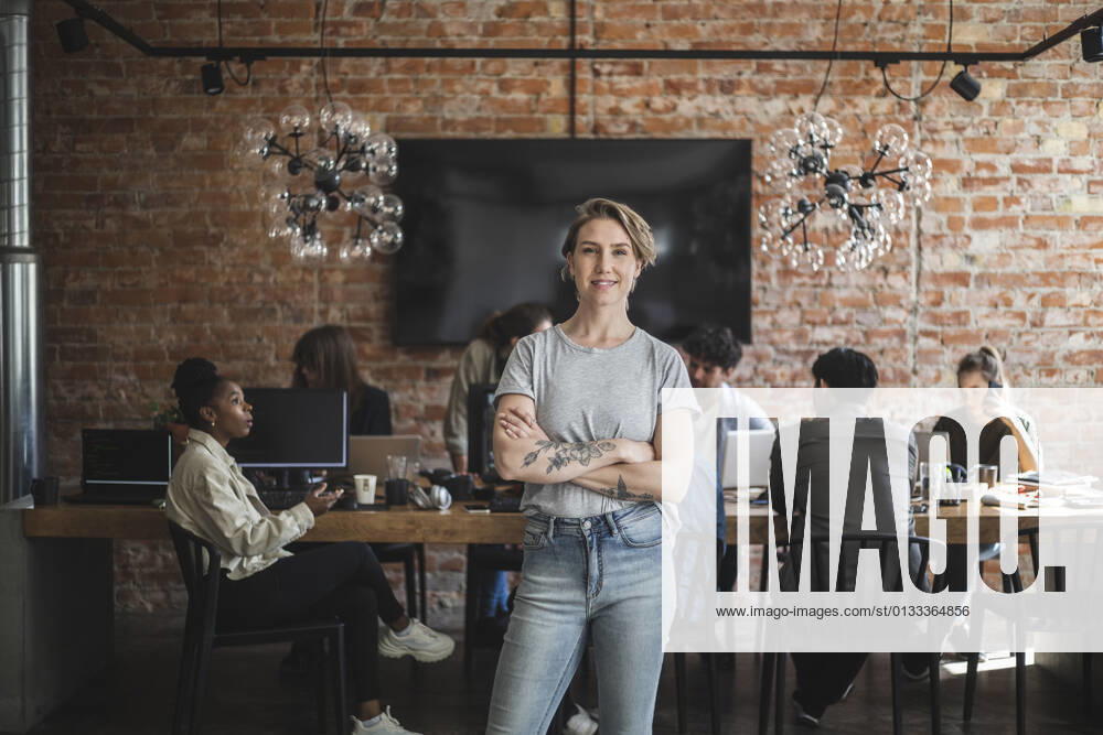 Female entrepreneur standing with arms crossed while colleagues working in creative office Sweden, Stockholm, model released, property released