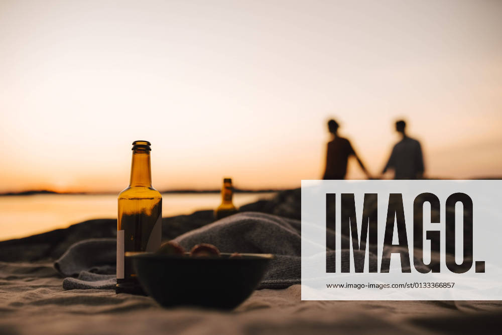 Beer bottle and bowl on picnic blanket while gay couple in background at lakeshore during sunset Sweden, Gothenburg, model released
