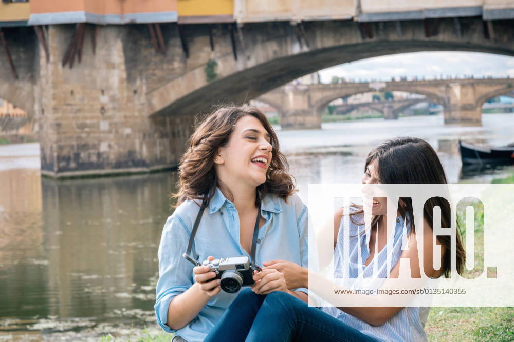 Lesbian couple sitting together looking at digital camera laughing, in front of Ponte Vecchio, Florence, Tuscany, Italy, 02.10.2015,