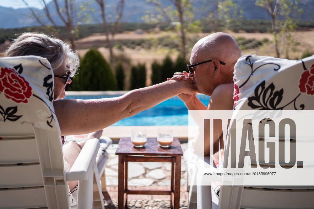 Back view of shirtless loving old husband kissing hand of wife while enjoying summer day on loungers on poolside, Model released JuanAlbertoRuiz_SeniorCoupleSwimmingPool_14.jpg