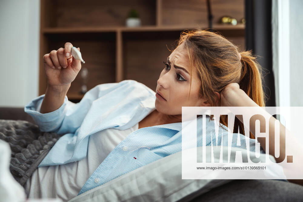 Oh No, Could It Be Flu? Young woman is lying sick at home couch and looking at thermometer.