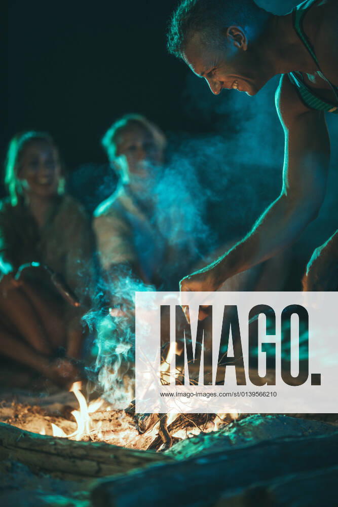 IMAGO Creative: Jahreszeit Frühling, Picknick Delicious Snacks The happy young smiling friends are sitting on the sandy seashore by the fire in the evening. They are frying sausages on stick.