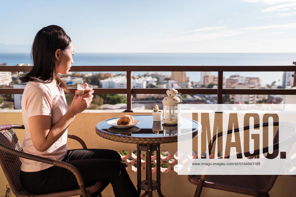 Happy Asian female with glass of hot drink in hand looking at camera while sitting on balcony at table with food during breakfast, Model released JuanAlbertoRuiz_BreakfastAtHome_1.jpg