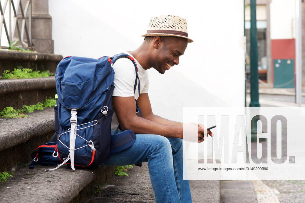 Side portrait of african american male tourist sitting on stairs with bag and mobile phone