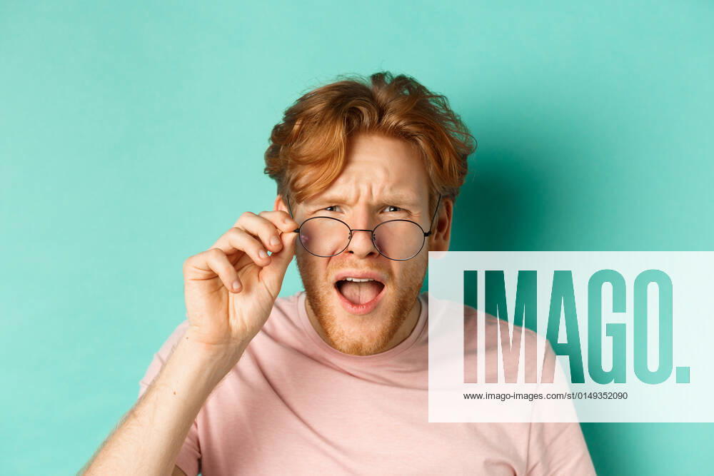 Close up portrait of redhead guy take-off glasses and looking confused at something strange, standing shocked over turquoise background Close up portrait of redhead guy take-off glasses and looking confused at something strange, standing shocked over turquoise background.