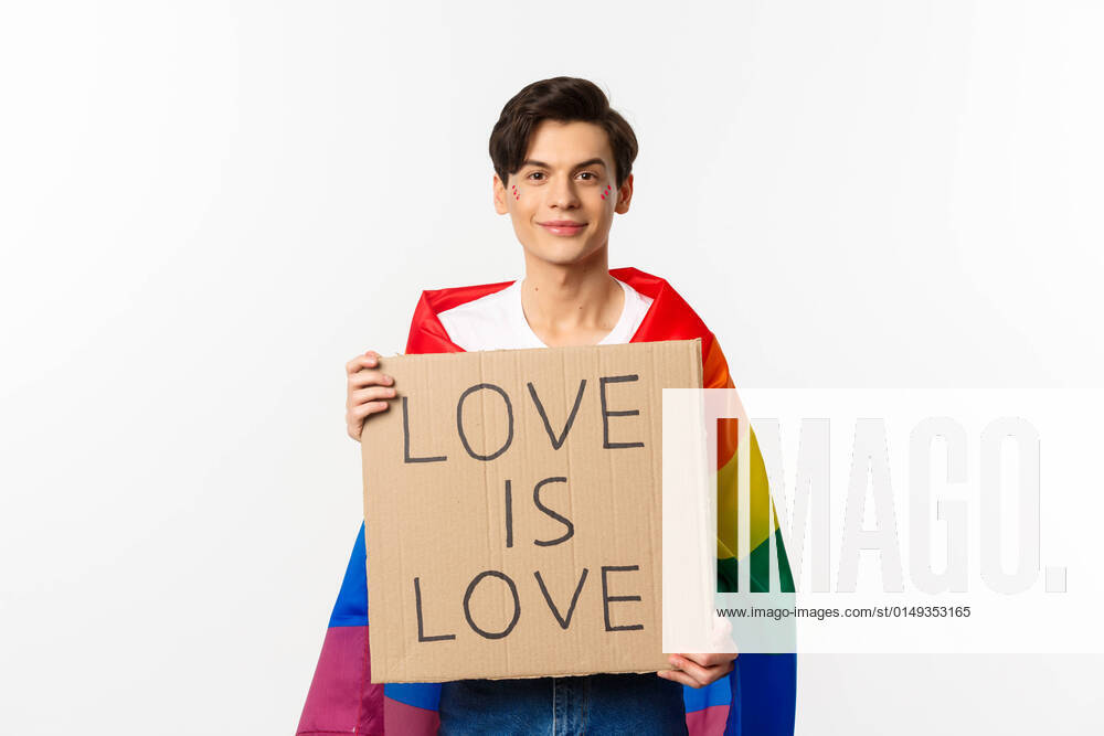 IMAGO Creative: Pride Month 2022, Engagement der Jugendlichen Smiling gay man activist holding sign love is love for lgbt pride parade, wearing Rainbow flag, standing over white background Smiling gay man activist holding sign love is love for lgbt pride parade, wearing Rainbow flag, standing over white background. Copy space