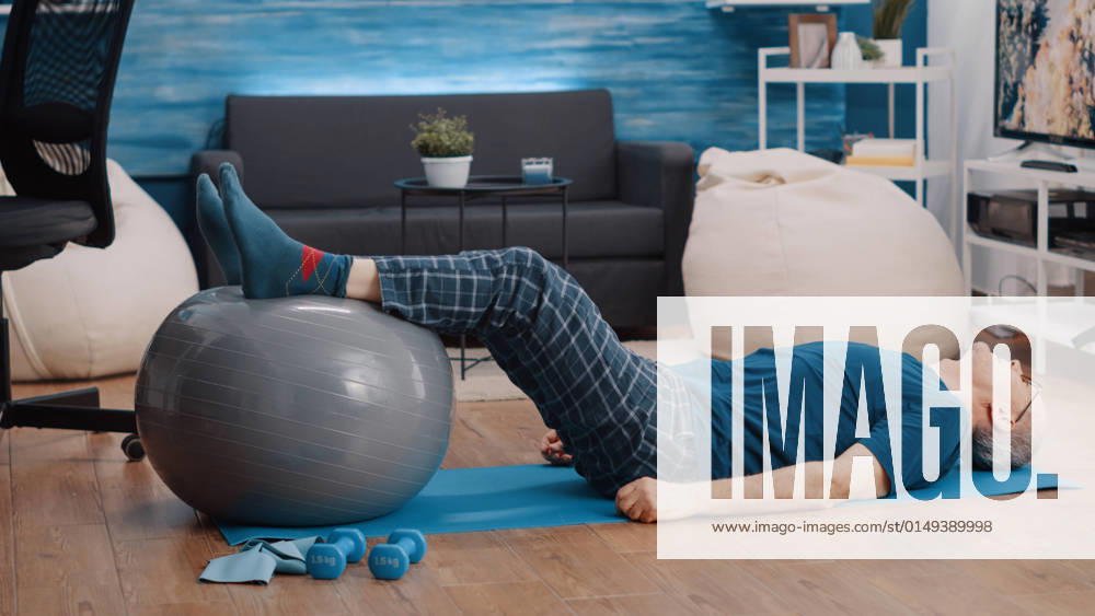 Retired person using fitness toning ball and lifting legs Retired person using fitness toning ball and lifting legs to stretch muscles. Aged man training and doing physical exercise while laying on yoga mat. Pensioner exercising with equipment