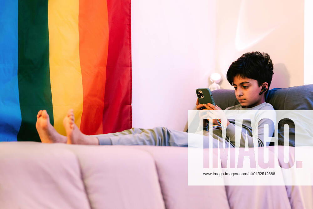 IMAGO Creative: Pride Month 2022, Engagement der Jugendlichen IMAGO Creative: Pride Month, Juni 2022 Full body side view of barefoot ethnic boy in wireless earbuds lying on bed and browsing cellphone near colorful LGBT flag HodeiUnzueta_YoungNonBinary_31.jpg