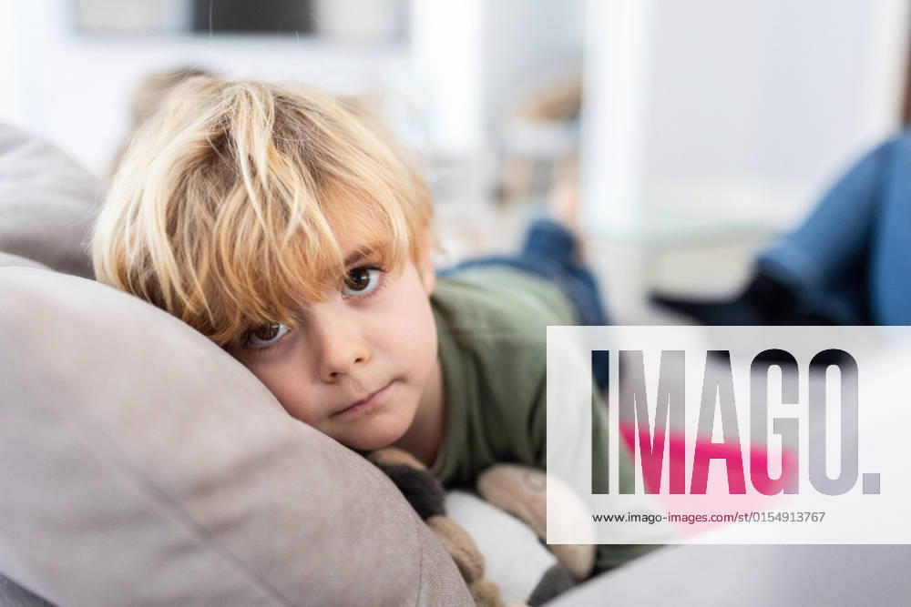 Unhappy boy with blond hair looking away while lying on comfortable gray sofa with toy in light living room on blurred background, Model released AngelSantamaria_DigitalDependency_10.jpg