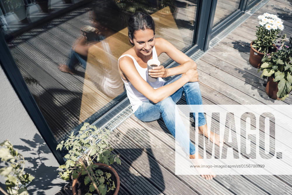 IMAGO Creative, Urban Lifestyle Happy woman with coffee cup sitting in balcony on sunny day model released, Symbolfoto property released, JOSEF07983
