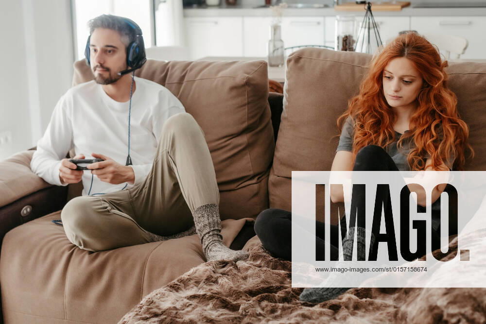 Full body of sad redhead female sitting on comfortable couch near boyfriend playing interesting videogame in light living room at home