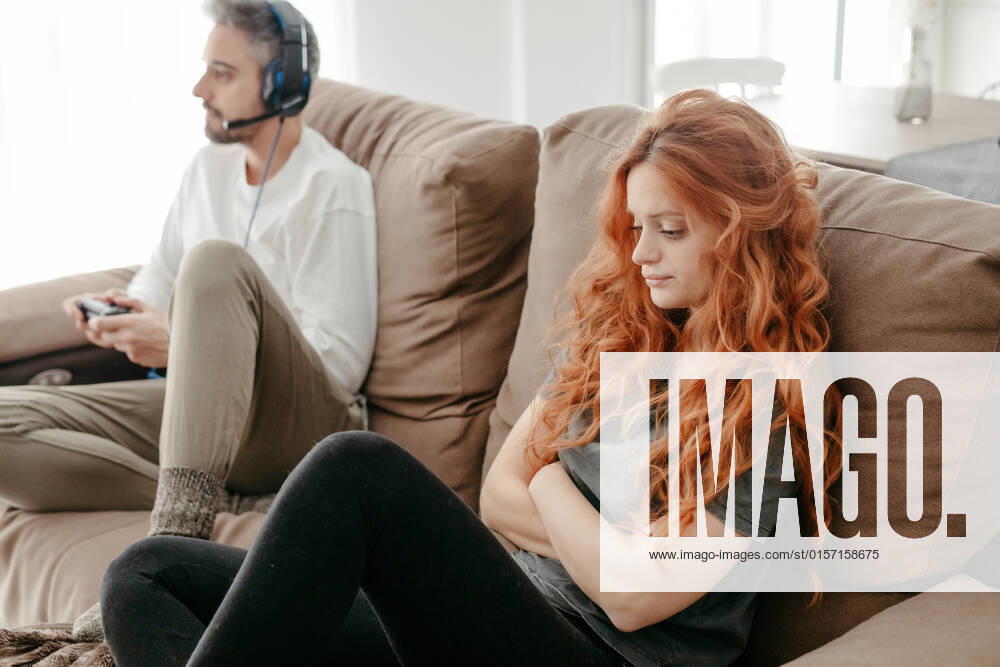 Full body of sad redhead female sitting on comfortable couch near boyfriend playing interesting videogame in light living room at home