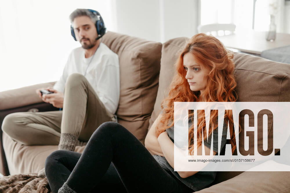 Full body of sad redhead female sitting on comfortable couch near boyfriend playing interesting videogame in light living room at home
