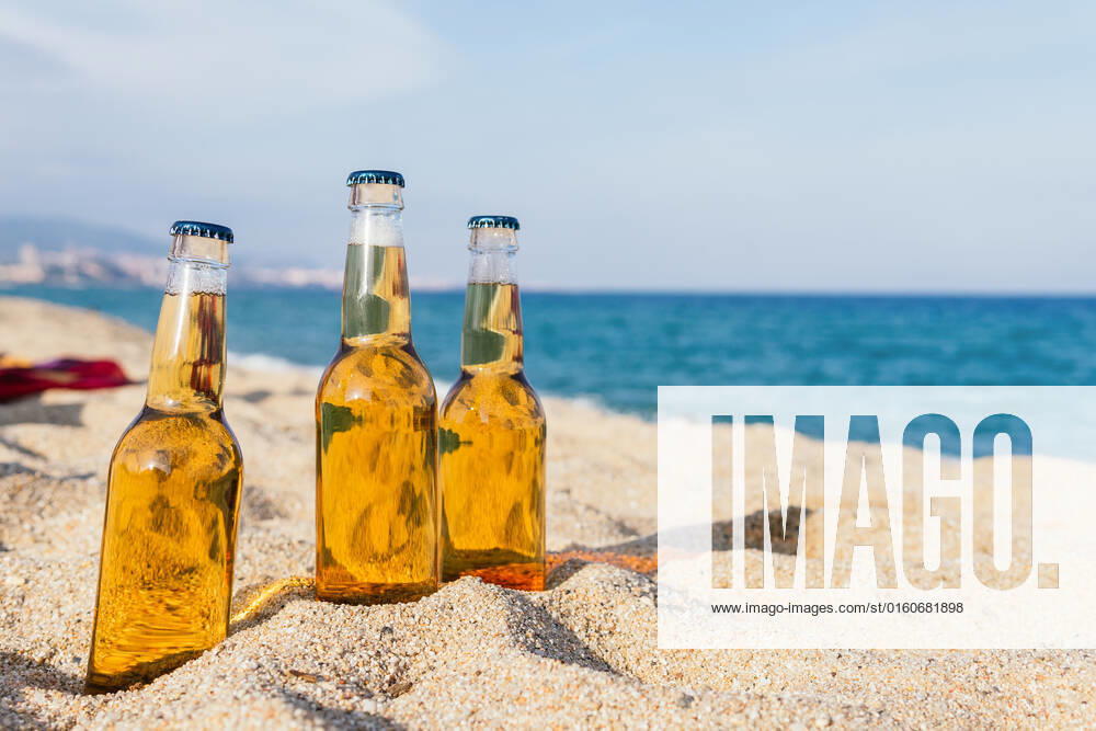 Glass bottles of cold beer placed on sand on blurred background of sea and blue sky on sunny day on resort