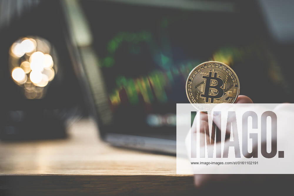 Hand hält eine Bitcoin Goldmünze BTC Cryptowährung vor einem Laptop Computer, Online Handel an der Börse mit Kurs Grafik
