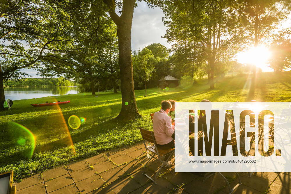 Menschen sitzen in der Abendsonne an einem Tisch einer Gartenwirtschaft und essen, Bayern, Deutschland. Gartenwirtschaft an einem Seeeinem kleinen See