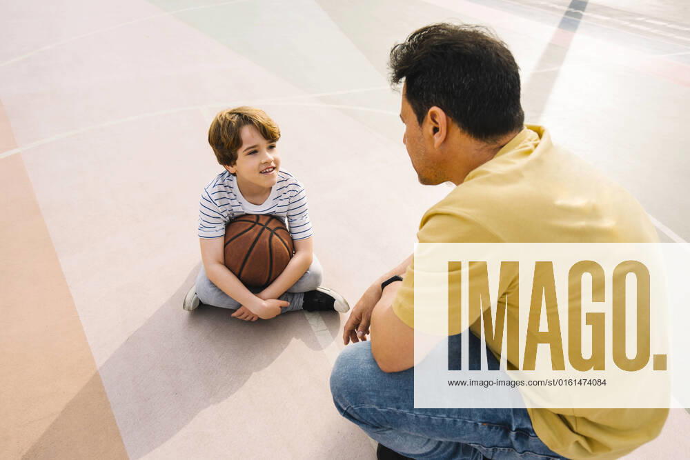 IMAGO Creative: 2022 Sport Favoriten IMAGO Creative: Lokalhelden - Jugendsportbildung Boy with basketball sitting in front of father at sports court on sunny day model released, Symbolfoto, MMPF00108