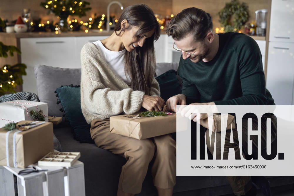 Smiling woman with man wrapping Christmas gift sitting on sofa at home model released, Symbolfoto property released, ABIF01717
