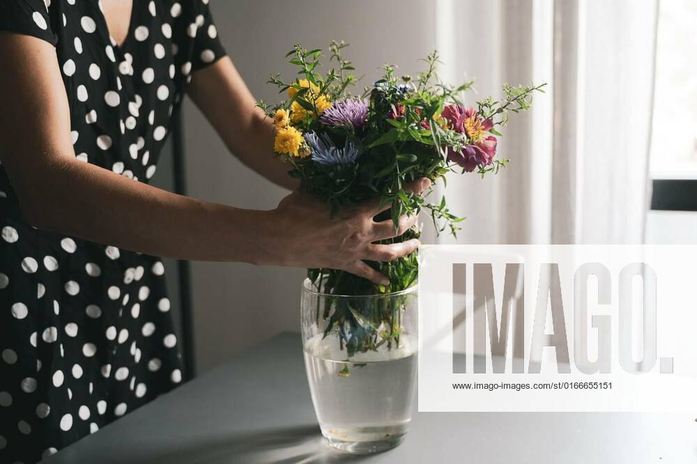Woman arranging a bouquet of flowers in a glass vase, young Caucasian woman with black dress and white polka dots arranging a bouquet of freshly picked up beautiful flowers in a glass vase on a grey table in natural day light model released, Symbolfoto , 17.03.2021,