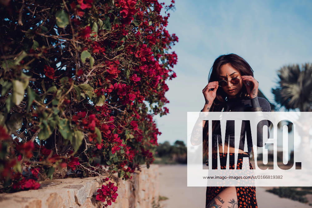 Confident stylish tattooed female with nose piercing and bindi touching sunglasses with closed eyes on the street against blurred shrub with vinous flowers, Model released RemediosPuerta_BohoBea_3