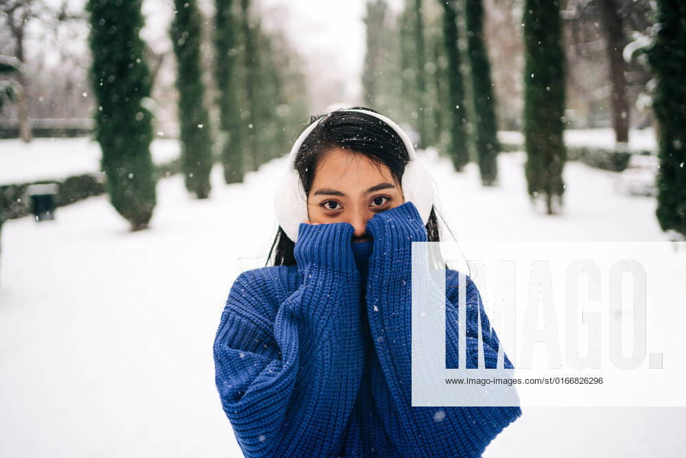 IMAGO Creative: Winter in der Stadt Young pleasant ethnic female in warm clothes covering face while looking at camera on snowy pathway in Madrid Spain, Model released JakeJakab_SnowyAfternoonShoot_14