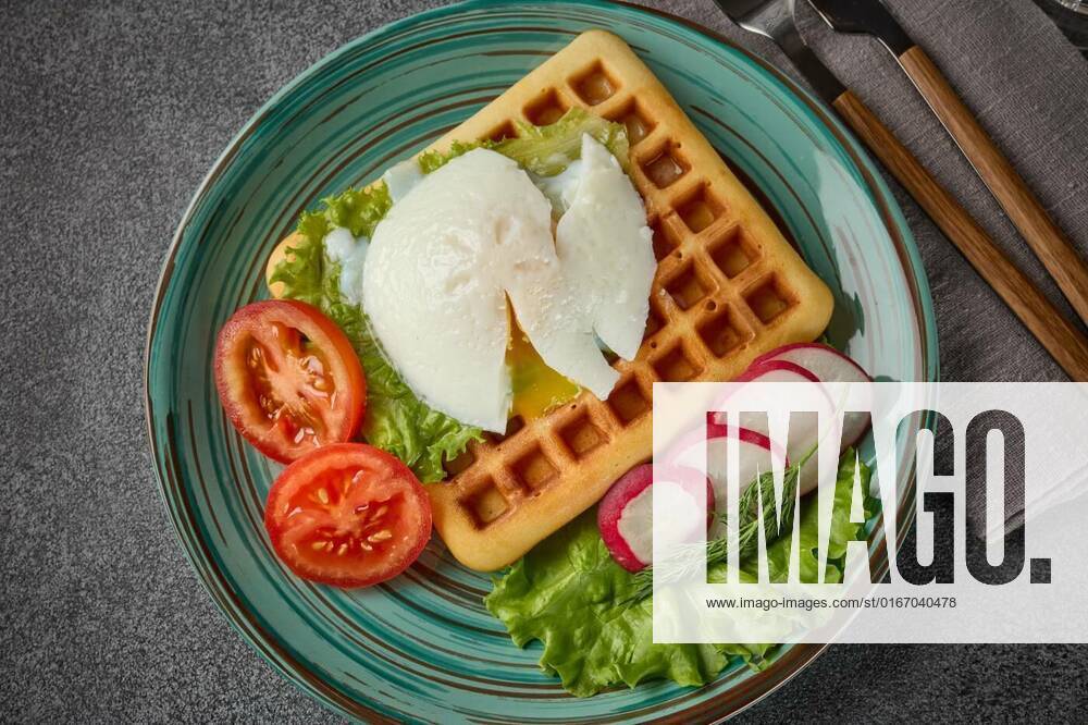 Tasty waffles with poached egg and vegetables on blue plate on grey background. Top view. Table setting, Delicious waffles with poached egg and fresh vegetables on blue glass plate on slate grey background. Top view. Serving food. Table setting, 20.06.2021,