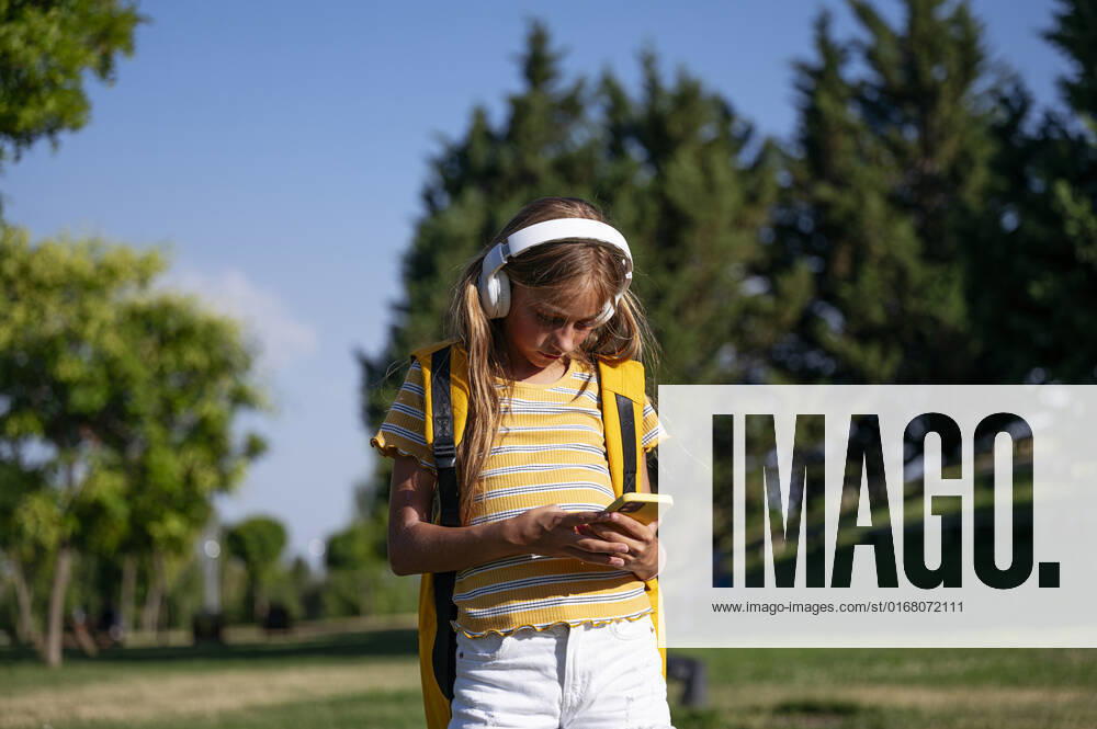 Focused teenage girl with backpack listening to music in wireless headphones while surfing cellphone in park on sunny summer day, Model released JoseLuisCarrascosa_ID441_16909_073