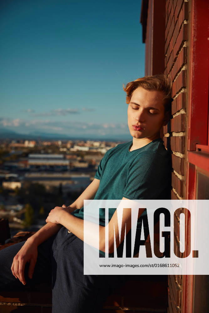 IMAGO Creative: 2022 Favoriten IMAGO Creative: St?dte im Wandel Cute young blond male resting with closed eyes sitting on edge of roof leaning against brick wall on sunny day, Model released CienXCienStudio_DancerBoy_70
