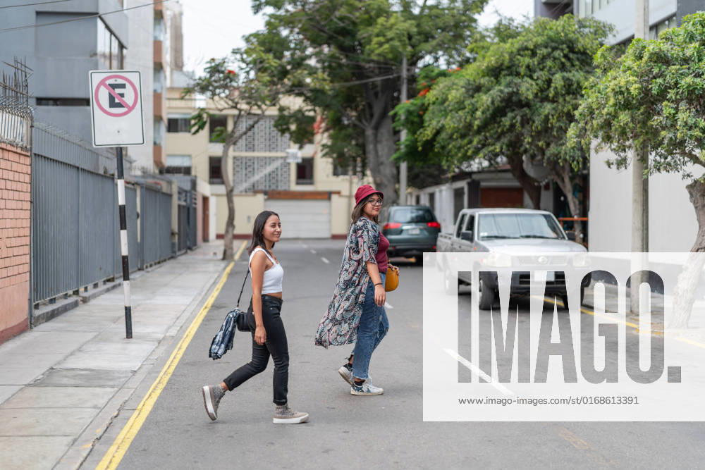 Full body of positive Peruvian female friends crossing the road in a street with parked vehicles and surrounded by trees and buildings, Model released GuillermoSpelucin_SaraiAndPaulaInLima_4