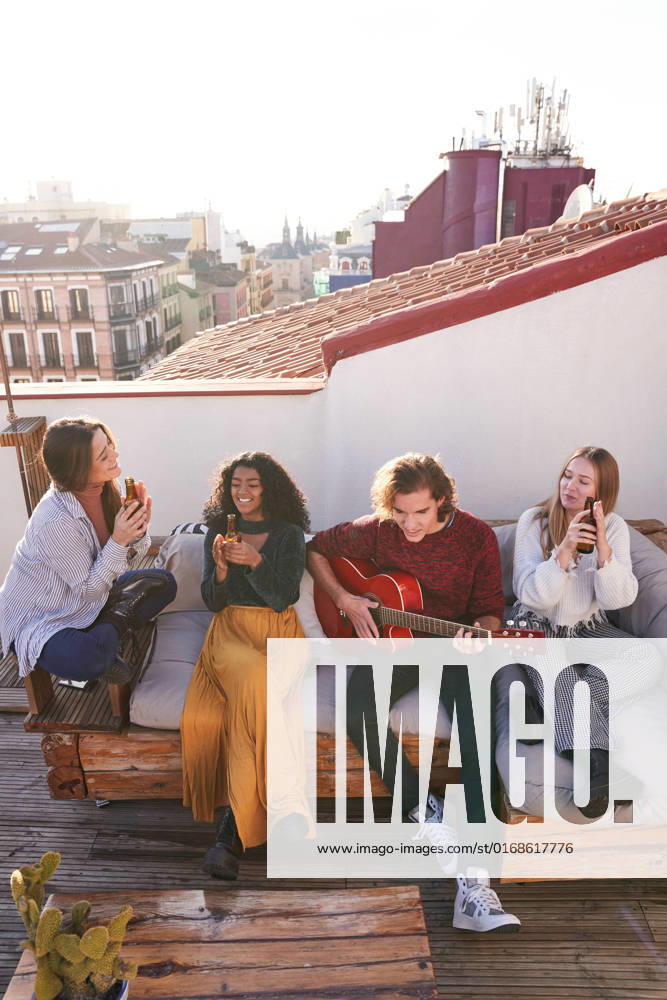 IMAGO Kreativ: Urbane Kultur - Sommer auf den D?chern Full length happy young multiracial friends in casual outfits playing guitar and enjoying beer while gathering together on comfy sofa on terrace, Model released, Property released ManuelRuiz_FlatmatesMusiciansTerrace_32