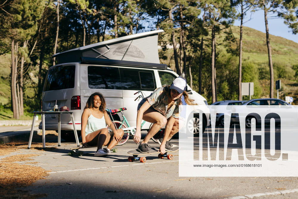 IMAGO Creative, Jahreszeit Sommer, Sommerferien Beautiful skater practicing riding skate board on street with background of woman sitting on skate near minivan in camping, Model released ManuelRuizAlba_CamperGirls_84