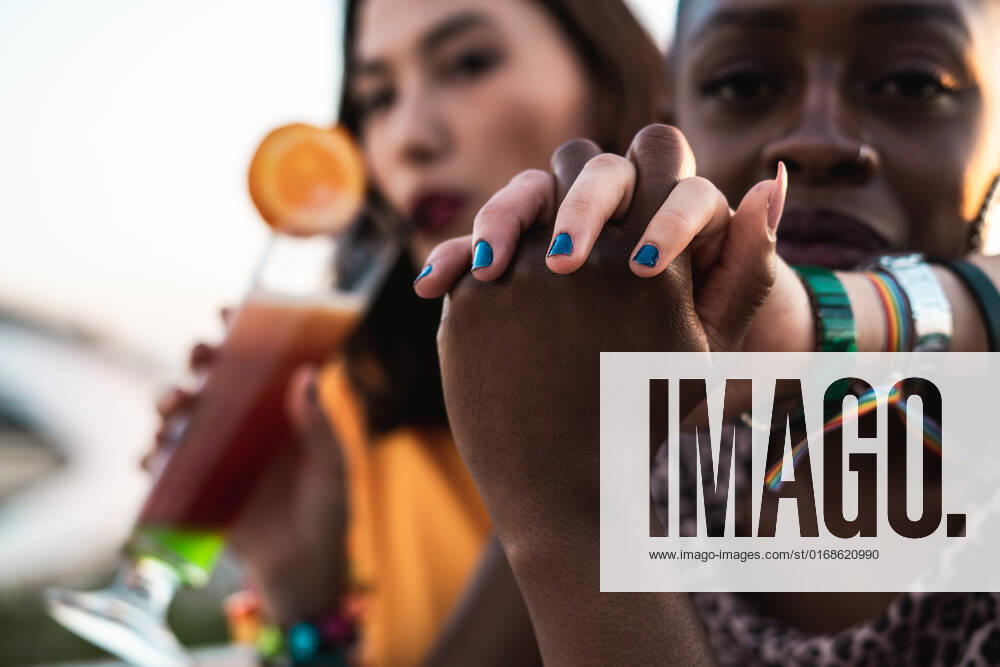 Stylish multiracial girlfriends sitting in open air cafe hugging and holding hands while enjoying refreshing drinks on blurred background, Model released RafaCortes_LesbianEthnicCouple_118