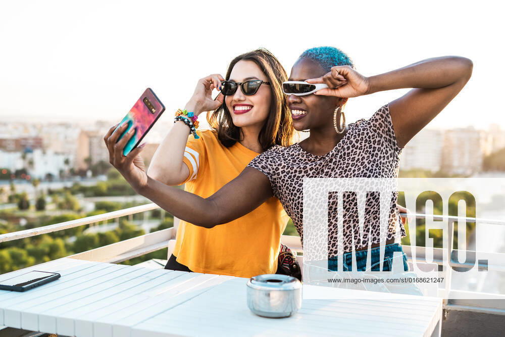 Cheerful multiracial girlfriends in trendy sunglasses smiling sitting at table taking selfie on urban background, Model released RafaCortes_LesbianEthnicCouple_96