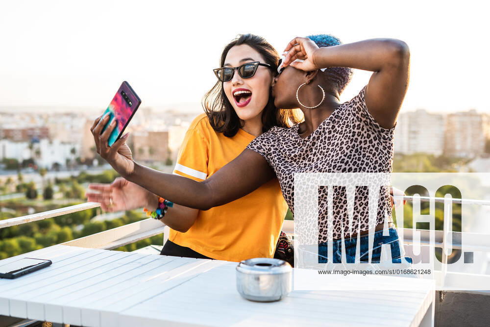 Cheerful multiracial girlfriends in trendy sunglasses smiling sitting at table taking selfie on urban background, Model released RafaCortes_LesbianEthnicCouple_97