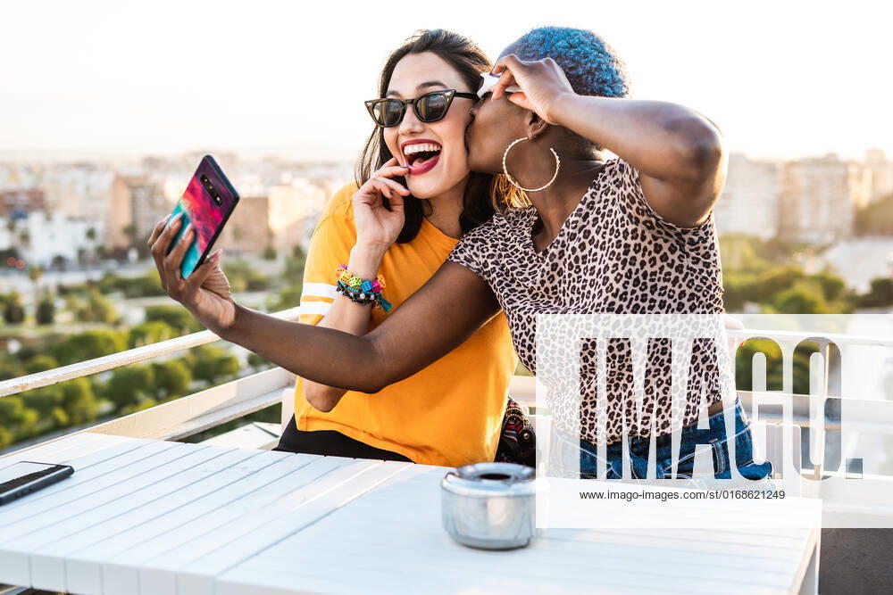 Cheerful multiracial girlfriends in trendy sunglasses smiling sitting at table taking selfie on urban background, Model released RafaCortes_LesbianEthnicCouple_98