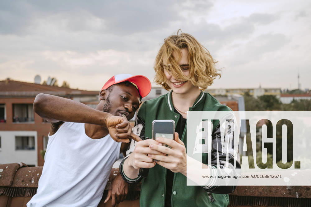 IMAGO Kreativ: Urbane Kultur - Sommer auf den Dächern IMAGO Creative: Business, Innovative Technologie-Lösungen Young man wearing hat pointing on smart phone held by male friend on rooftop Germany, Berlin, model released, property released