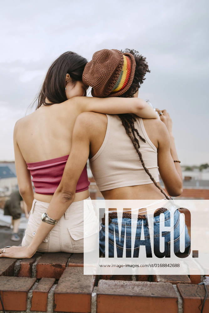 IMAGO Kreativ: Urbane Kultur - Sommer auf den Dächern Rear view of young woman embracing female friend wearing knit hat while sitting on rooftop Germany, Berlin, model released, property released