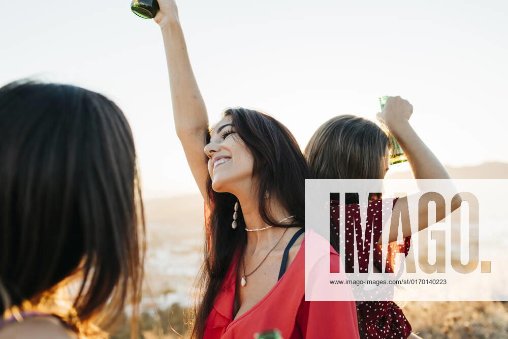 Delighted female friends in stylish outfits with bottles of alcohol beverages standing back to back while dancing on hilltop over town against cloudless sky in sunlight, Model released Lightsy_FriendsMountain_45