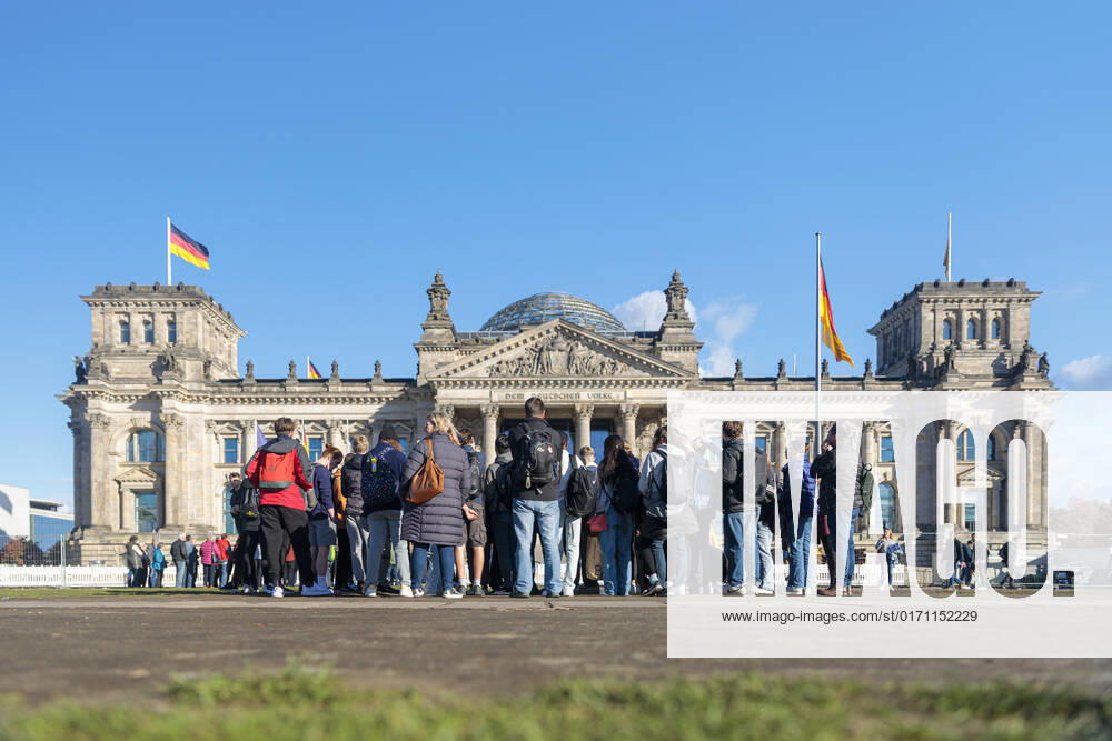 Symbolbild Deutscher Bundestag Deutschland, Berlin , Platz der Republik am 19.10.2022: Der Reichstagbau in der Herbstsonne mit blauem Himmel und weißen Wolken, wehender Deutschlandfahne. Im Vordergrund junge Menschen in Rückenansicht, die sich interessiert dem Reichstag zuwenden.