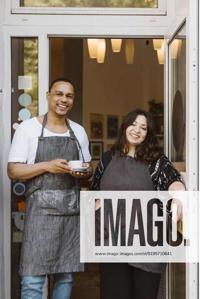 Portrait of happy multiracial male and female cafe owners standing at doorway