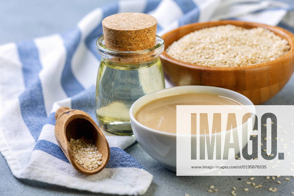 Bottled sesame oil, tahini and white sesame seeds. Cold pressed sesame oil from white sesame, tahini and sesame seeds in wooden bowl on grey textured background, selective focus.