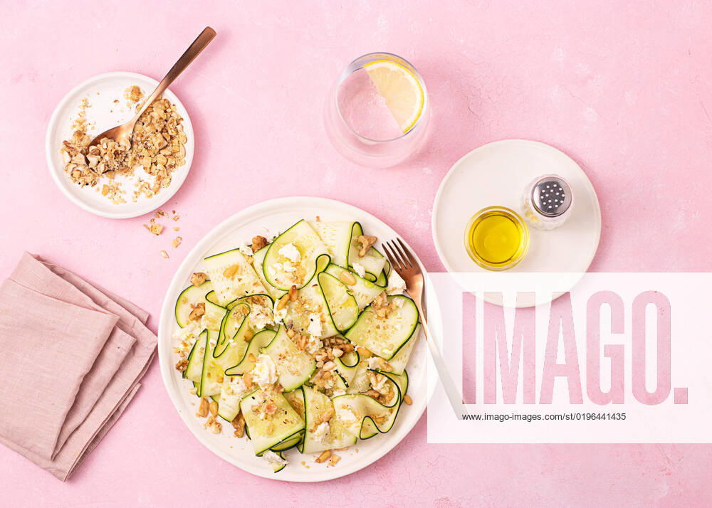 From above fresh salad of thin sliced ripe zucchini, feta cheese and pine nuts in white plate on table pink background OlgaVila_ID10586_320789_004