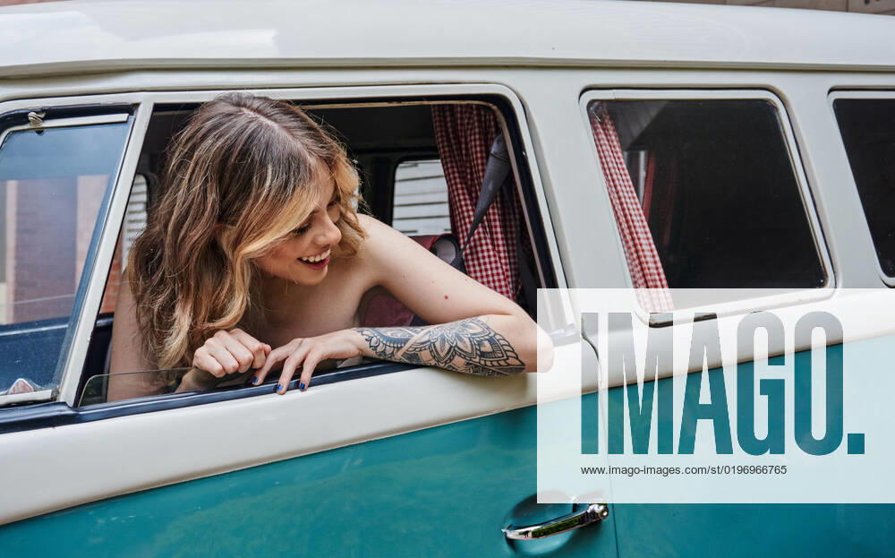 IMAGO Creative, Jahreszeit Sommer, Sommerferien Delighted young female driver with long wavy hair and tattoo on arm smiling and looking away while sitting in vintage van during holidays JoseDeJesusSaldana_ID10013_302921_038